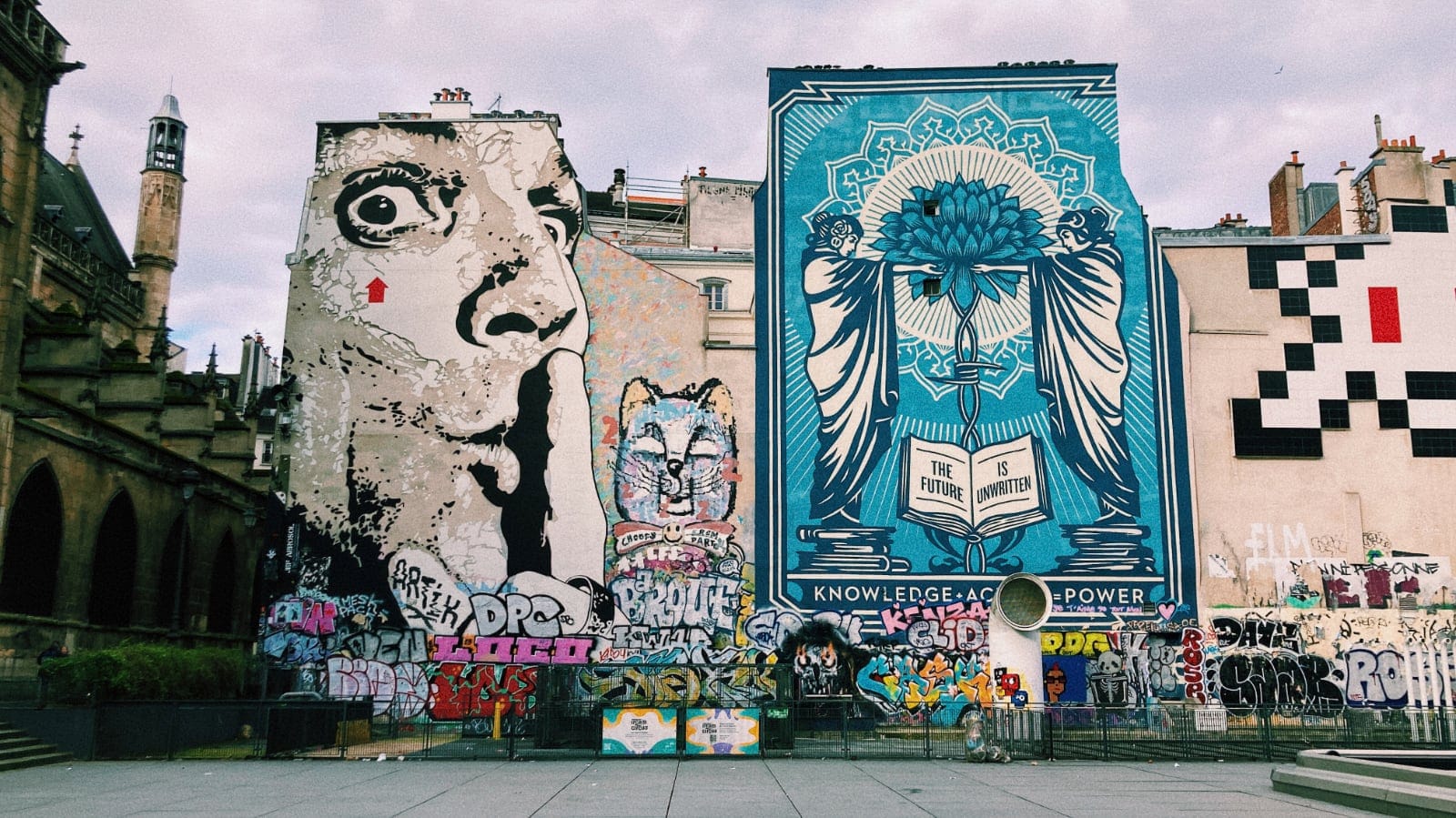 A photo of giant murals in Paris. On the right, there is one by Shepard Farey called "The Future is Unwritten" showing two women standing on a pile of books and holding a lotus flower